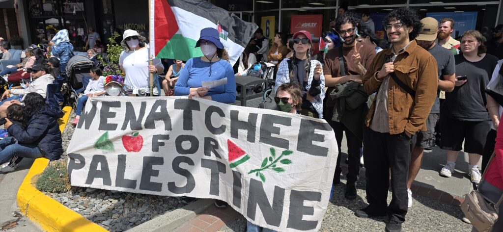Protesters from Wenatchee for Palestine at the Stemilt Grand Parade. They are holding a banner that says "Wenatchee for Palestine" and a large Palestinian flag, as well as some smaller Palestinian flags.